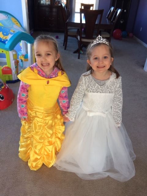 Alexandra pictured on the left and Michaela pictured on the right are dressed up in their dress up gowns holding hands.  Alexandra is wearing a yellow Belle ( Beauty and the Beast)  gown, and Michaela is wearing  a white wedding dress.